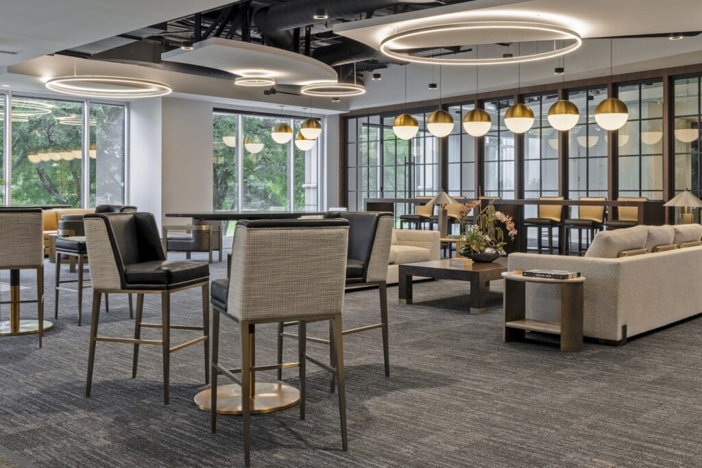 A bright, contemporary commercial office lounge featuring a plush gray sectional sofa and low-profile cream bouclé armchairs arranged around a round stone-top coffee table. In the background, floor-to-ceiling windows look out onto lush green trees, filling the space with natural light.